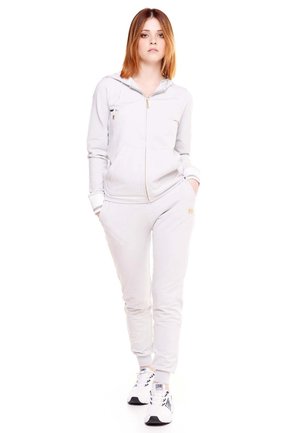 Une femme porte un sweat à capuche zippé gris clair et un pantalon de jogging assorti, avec des baskets blanches, sur un fond blanc uni.