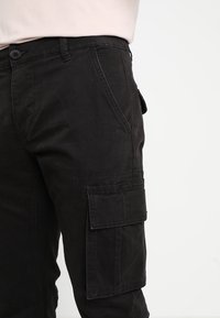 Czarne spodnie cargo wykonane z trwałego materiału, z boczną kieszenią, zapięciem na guzik i dopasowanym krojem. Struktura gładka w dotyku.