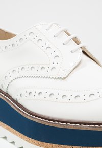 Chaussure brogue en cuir blanc avec perforations en forme d'aile, lacets blancs, semelle intermédiaire bleue et semelle plateforme texturée liège.