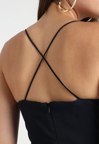 Robe noire avec des fines bretelles croisées à l'arrière et une fermeture éclair discrète, fabriquée dans un tissu lisse avec un design épuré.