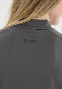 Tummanharmaa urheiluasema, jossa on korkea kaulus ja takana pieni musta "SLOPE" -logo. Sujuva kangasrakennus, istuva muotoilu.