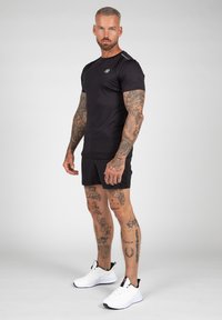 Zwart sport T-shirt met korte mouwen en logo, gecombineerd met zwarte shorts. Witte sneakers met zwarte accenten en patroon tattoo's op de benen.