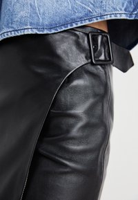 Pantaloni in pelle nera con una texture liscia, un dettaglio con fibbia laterale e una vestibilità aderente, parzialmente coperti da una maglietta in denim chiaro.