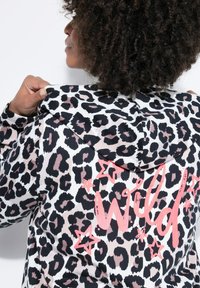Hættetrøje med leopardprint i hvid, sort og pink, med snøre i hætten og ordet "Wild" skrevet i pink skrift på ryggen med stjerneaccents.
