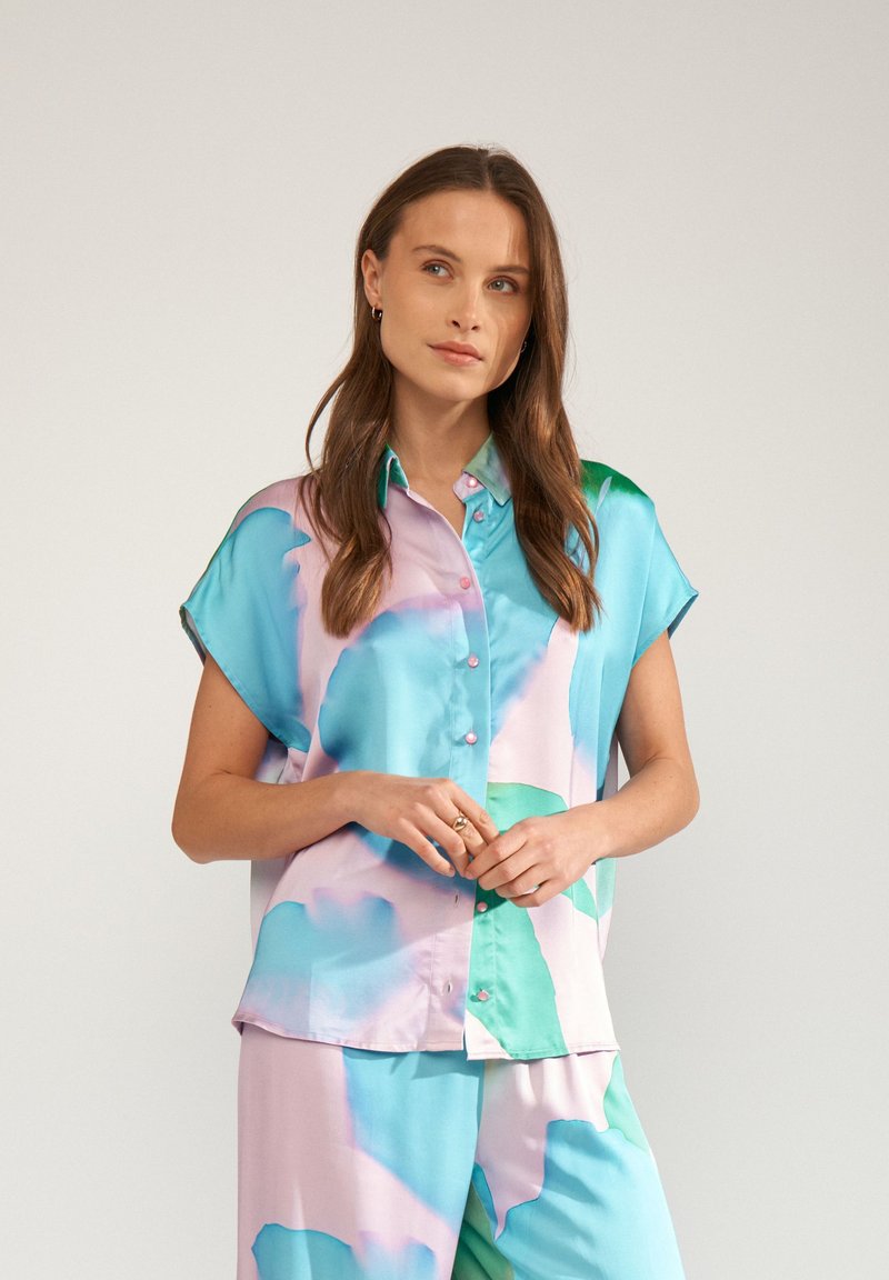 Femme aux longs cheveux bruns portant une chemise ample à manches courtes, boutonnée, de couleurs pastel bleu, vert et rose, avec un short assorti, debout devant un fond uni.