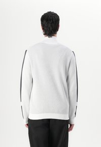 Svetlo sivi pulover z teksturiranim rebrastim vzorcem, z dodanimi temno sivimi stranskimi akcenti. Zapenjanje ovratnik, dolgi rokavi in sproščen kroj.