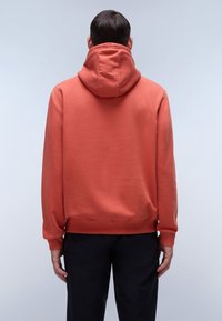 Felpa arancione con cappuccio, dal tessuto morbido, con polsini e orlo a coste, dotata di un grande cappuccio rilassato. Indossata con pantaloni neri.