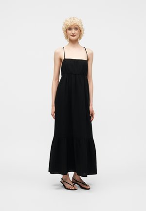 Γυναίκα που στέκεται με μαύρο μακρύ φόρεμα χωρίς μανίκια με λεπτές τιράντες και μαύρα σανδάλια με λουράκια πάνω σε απλό λευκό φόντο.