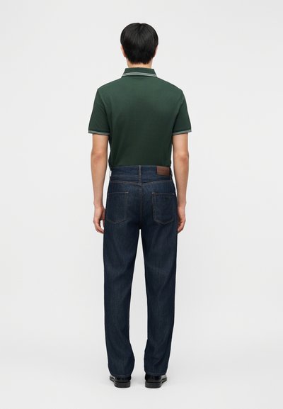 Άνδρας με πλάτη φοράει σκούρο πράσινο πόλο μπλουζάκι με κοντά μανίκια, σκούρο μπλε τζιν και μαύρα παπούτσια μπροστά σε απλό λευκό φόντο.