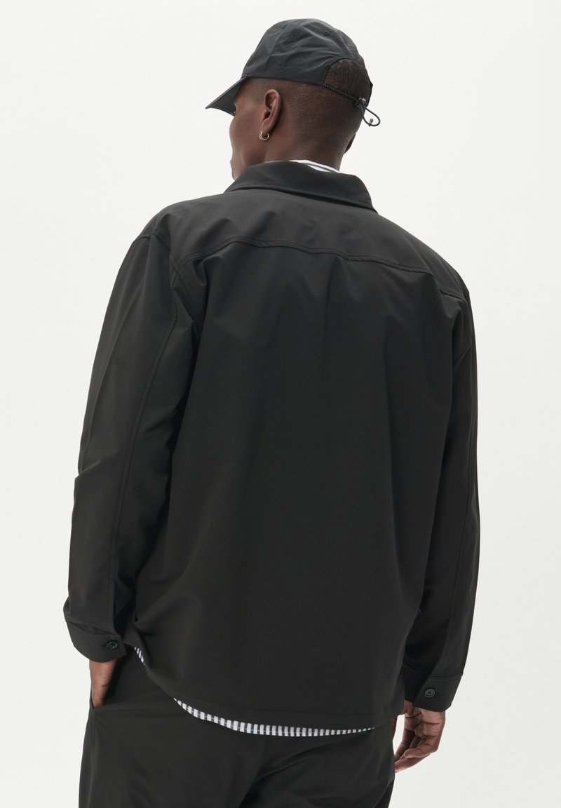 Czarna, lekka kurtka o gładkiej fakturze, z klasycznym kołnierzem, mankietami na guziki oraz delikatnie zaokrąglonym dołem. Noszona z pasującą czapką.