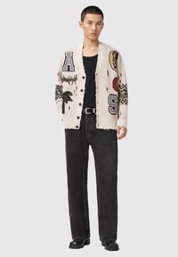 Πλεκτή ζακέτα σε κρεμ χρώμα με σχέδια, που περιλαμβάνει γράμματα και λουλουδένια μοτίβα. Συνδυάζεται με μαύρα τζιν και μαύρες μπότες. Κουμπώνει μπροστά με κουμπιά.