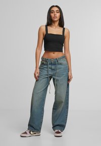 Žena stoji i nosi crni kratki top, široke isprane plave traperice s remenom i smešano-ružičaste patike.