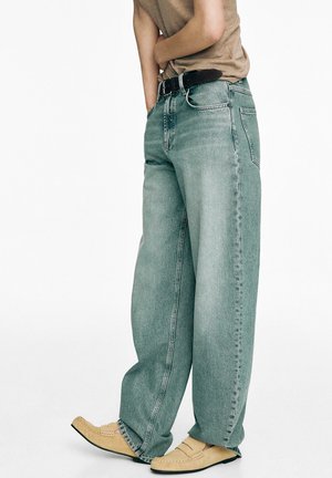 Personne portant un jean ample délavé, une chemise beige, une ceinture noire et des mocassins beiges, debout avec une main dans la poche devant un fond uni.