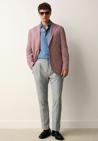 Blazer texturé rose clair sur une chemise en chambray bleu, associé à un pantalon à carreaux gris et des chaussures noires, des lunettes de soleil complètent le look.