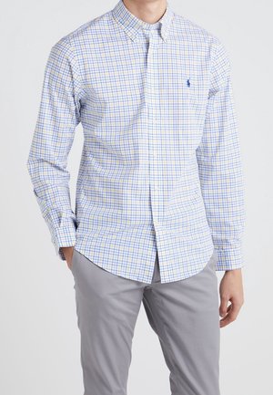 Mężczyzna w białej koszuli z guzikiem w niebiesko-żółtą kratę i szarych spodniach, stojący z rękami w kieszeniach.