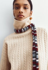 Kremowy sweter z warkoczowym splotem i wysokim ribbanym kołnierzem, podkreślony wielokolorowym szalem w szarych, czerwonych i różowych odcieniach.