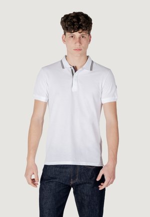 Polo shirt bianco con apertura a due bottoni, dettagli del colletto a righe nere e grigie, maniche corte e texture di cotone liscia. Indossato con jeans scuri.