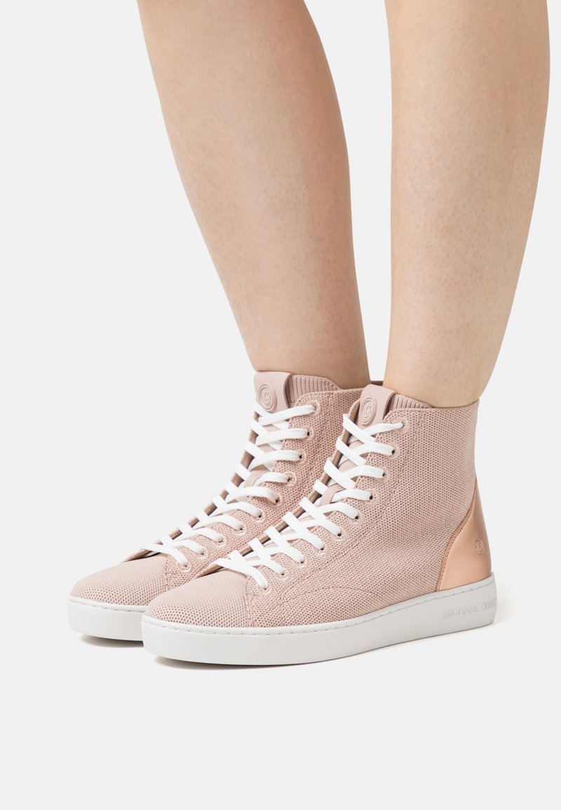 Baskets montantes en maille rose clair avec des lacets blancs et des semelles en caoutchouc, portées pieds nus sur un fond uni.
