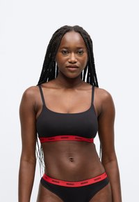 Čierny bralet s tenkými ramienkami a červenou elastickou páskou so slovom „HUGO“ na hrudníku. Zodpovedajúce čierne nohavičky s podobným dizajnom pásky.