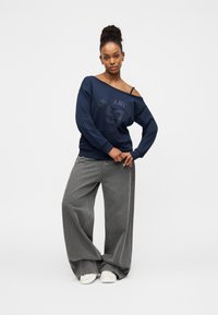 Μπλε σκούρο μπλουζάκι φούτερ με φθαρμένο γραφικό εκτύπωμα, συνδυασμένο με φαρδιά γκρι τζιν παντελόνια και λευκά αθλητικά παπούτσια. Το μοντέλο στέκεται σε χαλαρή στάση.