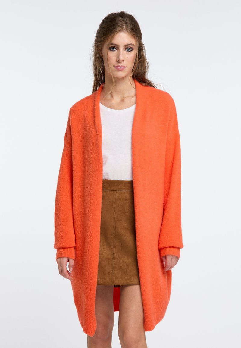 Översized orange stickad cardigan med öppen front, långa ärmar och ribbad mudd. Kombinerad med en ljus topp och kjol i brunt mocka.