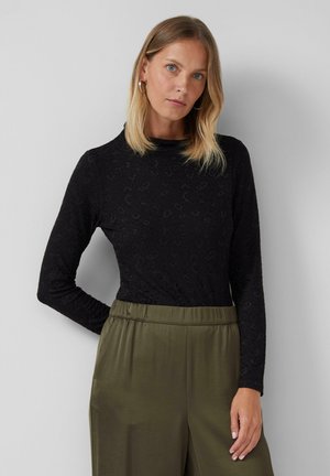 Femme aux cheveux blonds portant un haut à manches longues noir à motifs et un pantalon taille haute vert olive devant un fond uni.