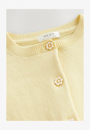 Cárdigan de punto amarillo con cuello redondo, que presenta dos botones en forma de flor blanca y la etiqueta "NEXT" cosida en su interior.