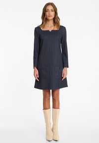 Frau in einem langärmligen, dunkelgrauen Nadelstreifenkleid mit cuadratischem Ausschnitt und vorderen Taschen, kombiniert mit beigen knielangen Stiefeln, aufrecht stehend.