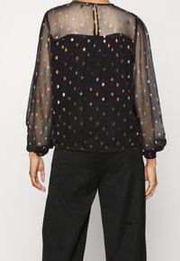 Blusa nera trasparente con maniche lunghe, caratterizzata da un motivo a pois color rame, scollatura arrotondata sul retro con chiusura a bottone e polsini arricciati.