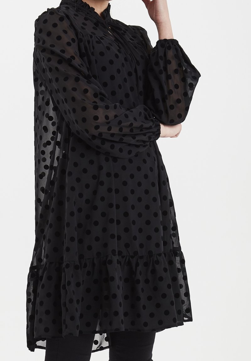 Musta pilkullinen mekko ohuesta kankaasta, jossa on röyhelöreuna, pitkät puffihihat ja mandariinikaulus kerätyillä yksityiskohdilla.