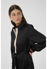 Rochie camasa neagra din satin cu un plastron de nasturi contrastant in crema, maneci lungi si talie legata. Textura neteda si design clasic al gulerului.