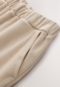Pantalon en velours côtelé beige avec taille élastique et poche latérale fendue, montrant la texture détaillée du tissu et les coutures.