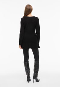 Zwarte tuniek met lange mouwen en een high-low zoom, gecombineerd met zwarte nauwsluitende legging en glanzende zwarte laarzen met hak tot aan de knie. Soepele textuur.