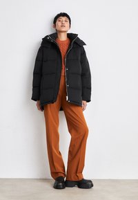 Svart puffjakke med hette, glidelås og kviltet design. Kombinert med en oransje strikkegenser og vide oransje bukser. Svart platåsko.