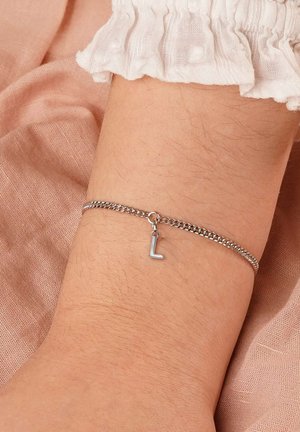 Silver chain bracelet with a dangling letter "L" charm on a wrist, resting on soft pink fabric and white ruffled fabric sleeve.