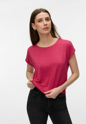 Kvinde med glat brunt hår iført små øreringe i form af øreringe, en kortærmet fuchsia top og sorte jeans, der holder i nederste kant af sin skjorte.
