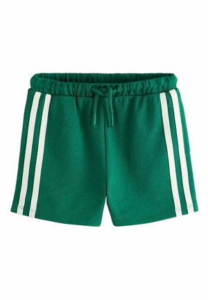 Småbarn iført hvide sneakers med velcro, hvide sokker, grønne shorts med hvide striber og en hvid t-shirt med en banangrafik og tekst.