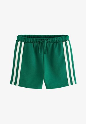 Dijete koje nosi bijele tenisice s čičak trakom, bijele čarape, zelene kratke hlače s bijelim prugama i bijelu majicu s grafikom banane i tekstom.