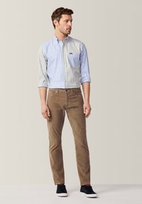 Gestreiftes Hemd in Hellblau und Weiß, braune Cordhosen und schwarze Sneaker mit weißen Sohlen, stehend vor einem neutralen Hintergrund.