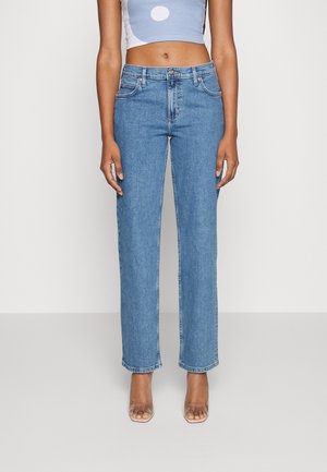 Persona que lleva vaqueros azules de talle alto y pierna recta, un top corto sin mangas con diseño circular y sandalias de tacón transparentes, de pie sobre un suelo gris.