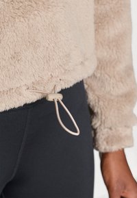 Pull en polaire fuzzy beige avec un cordon de serrage réglable à l'ourlet. Porté sur des leggings noirs, mettant en avant une texture douce et un design décontracté.