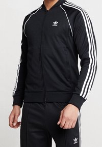 Svart zip-up jacka med vita ränder på ärmarna, tillverkad av ett slätt tyg. Har ribbade ärmslut och krage, med en liten logotyp på bröstet.