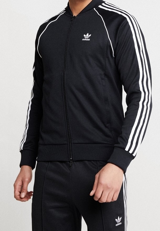 Svart zip-up jacka med vita ränder på ärmarna, tillverkad av ett slätt tyg. Har ribbade ärmslut och krage, med en liten logotyp på bröstet.