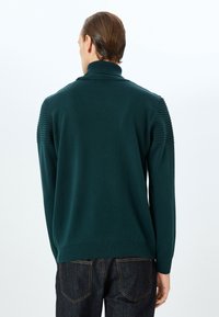 Mörkgrön tröja med hög krage, ribbad detalj vid axlarna och långa ärmar, tillverkad av ett mjukt stickat material. Matchas med mörka jeans.