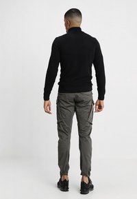 Zwarte gebreide coltrui met een textuurpatroon, gecombineerd met olijfgroene cargobroek met zijzakken en elastische manchetten. Zwarte sneakers maken de look af.