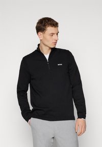 Black pullover sweater with a half-zip collar, ribbed texture, and white "BOSS" logo on the chest, paired with light gray trousers.