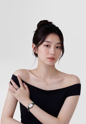 Mladá žena s tmavými vlasy sepnutými do drdolu, oblečená v černém topu s odhalenými rameny, se stříbrným náhrdelníkem, hodinkami se zeleným páskem a stříbrnými prsteny na ruce.