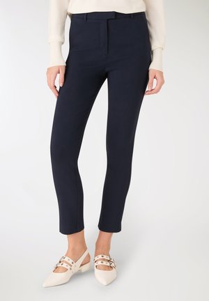 Pantaloni blu navy con una vestibilità su misura, fronte piatto e texture liscia. Indossati con scarpe slingback color crema con cinturini decorativi e dettagli in metallo.