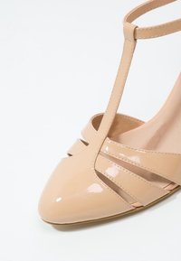 Béžová lakovaná kožená vysoká bota na podpatku se špičatou špičkou, designem T-strap a výřezovými detaily po stranách. Hladká textura.