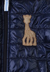 Giacca imbottita blu navy con chiusura a zip, caratterizzata da un logo di giraffa ricamato color beige. Texture liscia con pannelli cuciti e una tasca laterale.